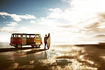 Das Foto zeich einen Surfer neben seinem Auto, einem VW Bus, an einem Strand 